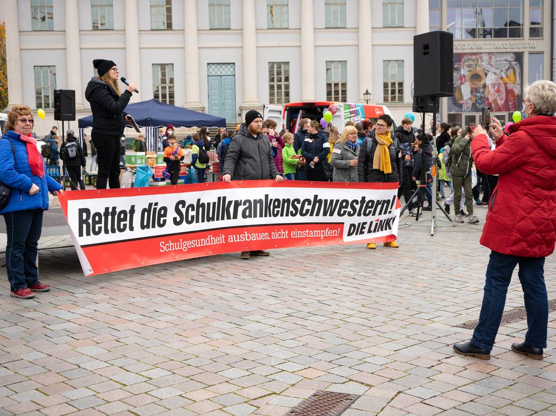 Anja Mayer steht mit einem Mikrofon in den Händen auf einem Podest, davor stehen mehrere Personen mit einem Transparent mit der Aufschrift "Rettet die Schulkrankenschwestern! DIE LINKE."